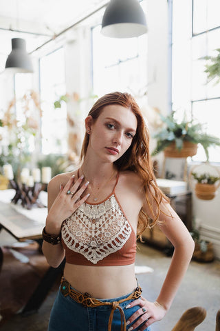 A woman with long reddish hair stands in a bright, airy room with large windows and hanging plants. She wears the Reina Crochet Lace High Neck Bralette by Leto Collection and jeans, with one hand touching her chest and the other resting on her hip, looking directly at the camera.