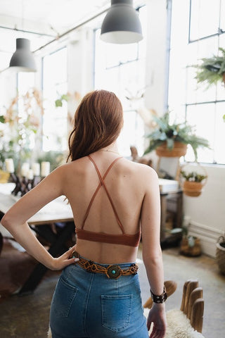 A person with long red hair stands with their back to the camera in a well-lit room adorned with hanging plants. They wear the Reina Crochet Lace High Neck Bralette from Leto Collection, paired with high-waisted blue jeans and a decorative belt.