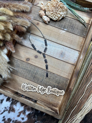 A rustic tray showcases the Josette Hand Craft Stone Y Necklace from Western Edge Boutique, which features a burnished silver design with rectangular black stones. Surrounding the necklace are dried florals, a faux flower, and a label that reads "Western Edge Boutique." The background consists of wooden textures and animal print fabric. The necklace measures 24″ with an additional 3″ extension.