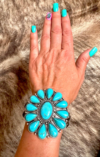 A hand with turquoise-colored nails rests on a fur background, showcasing the Raleigh Turquoise Stretch Bracelet from Western Edge Boutique, an ornate and antique silver plate bracelet adorned with multiple turquoise stones arranged in oval and teardrop patterns.