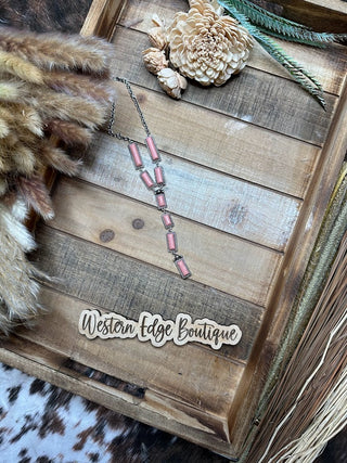 A rustic wooden tray holds the Olette Hand Craft Stone Y Necklace featuring rectangular salmon pink stones arranged vertically, framed by burnished silver accents. Beside it are decorative elements including a cream-colored flower, fur accents, and a feather. A sign reading "Western Edge Boutique" is placed at the bottom.