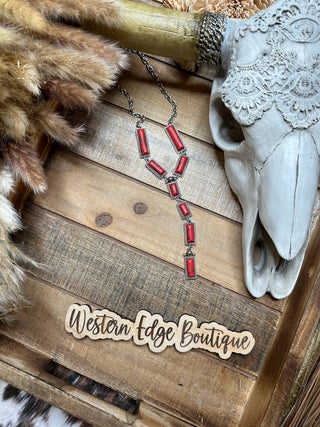 A rustic display features the Rosette Hand Craft Stone Y Necklace from Western Edge Boutique, adorned with burnished silver and red rectangular stones, measuring 24″ + 3″ EXT, arranged in a zigzag pattern. Surrounding the necklace are a lace-decorated skull, dried grasses, and a wooden plaque reading "Western Edge Boutique" on a wood-textured background.