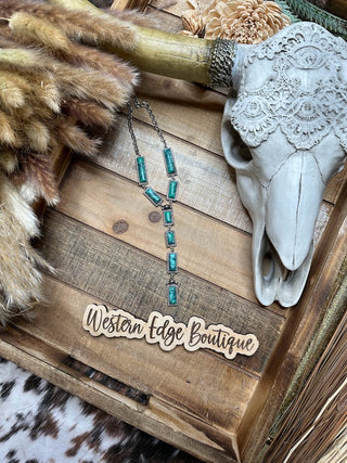 The Nanette Hand Craft Stone Y Necklace, featuring turquoise stones, a 24″ + 3″ extension, and burnished silver accents, is elegantly displayed on a wooden tray with a rustic, Western theme. The tray also holds a faux animal skull adorned with lace, a bundle of dried grass, and a sign reading "Western Edge Boutique." The setting is completed with patterned fabric in the background.