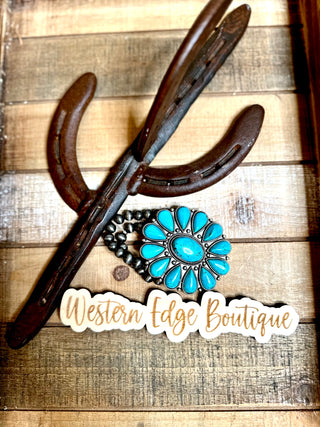 A decorative arrangement on a wooden surface featuring crossed horse shoes, a turquoise-stone bracelet with floral design, and a "Western Edge Boutique" sign written in cursive. The Raleigh Turquoise Stretch Bracelet by Western Edge Boutique adds an extra touch of charm to this rustic display.