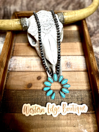 The Houston Turquoise Squash Blossom Necklace, beautifully arranged on a faux cow skull with horns, rests in a wooden tray. A rustic sign below reads "Western Edge Boutique" in cursive text. The background features a fur-like textured surface, perfectly highlighting the unique jewelry display.