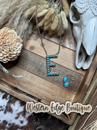 An Eliza "E" Letter Turquoise Necklace Earring Set is displayed on a wooden surface. An arrangement of dried wheat, a white decorative flower, and an animal skull are placed around the jewelry. Western Edge Boutique is written at the bottom.