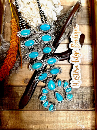 A decorative wooden tray holds turquoise jewelry pieces, including the stunning Quinlynn Squash Blossom Necklace adorned with silver accents, earrings, and a bracelet. Surrounding the jewelry is an assortment of earthy items such as an orange beaded decoration, dried flowers, feathers, and a sticker featuring the "Western Edge Boutique" brand.