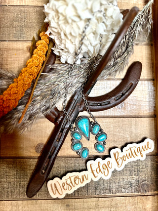 A rustic decor setup on a wooden surface features a horseshoe, white flower, orange and gray dried grass, and the Hazel Turquoise Squash Blossom Necklace. The scene includes a cutout text stating "Western Edge Boutique.