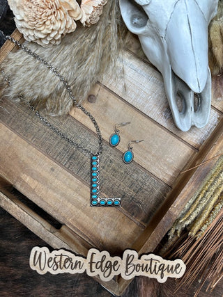 A rustic setup showcases the Lucy "L" Letter Turquoise Necklace Earring Set on a wooden surface, featuring a turquoise-accented "L" necklace paired with matching turquoise stone earrings. A decorative skull and straw elements complement the jewelry, and the text "Western Edge Boutique" is displayed prominently at the bottom.