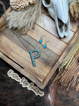 A wooden tray holds the Polly "P" Letter Turquoise Necklace Earring Set with brushed silver accents. On the tray lies a skull decoration and a small broom. A Western Edge Boutique sign and fur pelt are visible in the background.