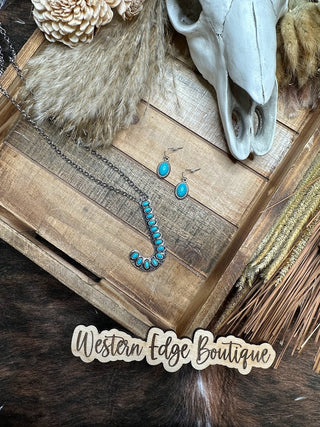 A rustic wooden tray showcases the Jayne "J" Letter Turquoise Necklace Earring Set, featuring a turquoise-studded pendant shaped like the letter 'J' and matching earrings. The tray is decorated with dried flowers, a skull, and other rustic elements. The caption reads "Western Edge Boutique – Curated by Ms. Jayne.