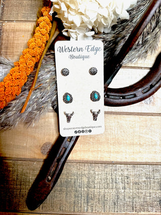 The Hawken 3 Pair Western Stud Earring Set from Western Edge Boutique is showcased on a card. The set comprises three pairs of earrings: one featuring turquoise stone accents and bull head designs, another with intricate burnished silver patterns, and an additional pair with unique western flair. The card rests on a rustic wooden surface adorned with decorative elements.