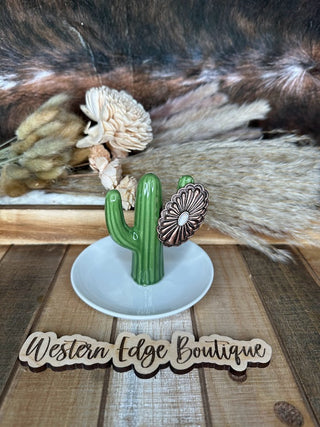 An Elsie Concho Cuff Ring from Western Edge Boutique is elegantly displayed on a ceramic green cactus ring holder, which features a dainty white daisy. The setup includes a white dish and dried flowers in the background, set against a rustic brown and white fur backdrop. A wooden sign with burnished copper accents prominently features the brand name, "Western Edge Boutique.