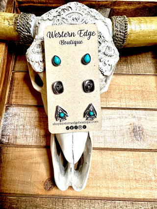 A decorative cow skull adorned with patterned fabric displays a cardboard backing with the Loper 3 Pair Western Stud Earring Set. The earrings feature silver tones with turquoise accents. The skull is mounted on a wooden wall, showcasing the "Western Edge Boutique" brand.