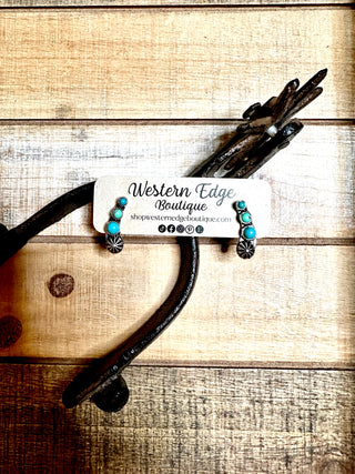 The Miley Turquoise Stud Concho Earrings from Western Edge Boutique are showcased on a rustic wooden surface, partially propped up by an old metal horseshoe. A card displaying the boutique's information accompanies the earrings.