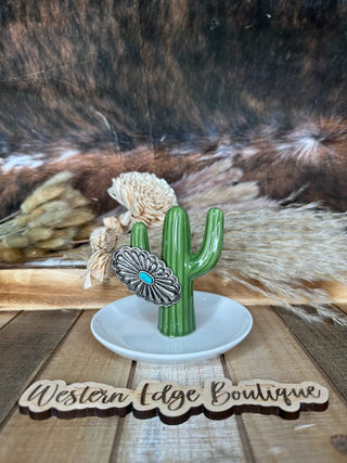 A decorative ceramic item shaped like a green cactus sits on a white dish. Adorning the cactus is the Lindlee Concho Cuff Ring by Western Edge Boutique, featuring a burnished silver, flower-shaped design with a turquoise-colored stone. The background includes dried grasses and a wooden sign reading "Western Edge Boutique.