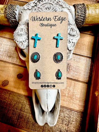 A product display from Western Edge Boutique showcases the Rogue 3 Pair Western Stud Earring Set on a rustic wooden background. This set includes earrings with cross, oval, and teardrop designs, each accentuated with turquoise stones and framed in burnished silver, elegantly mounted on a decorative skull.