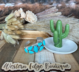 A wooden surface displays the Lottie Turquoise Cuff Bracelet, a green ceramic cactus on a white dish, and a bouquet of dried flowers. A sign reading "Western Edge Boutique" lies at the bottom. Set against wood and fur textures, the scene features a flexible cuff bracelet adorned with natural stones.