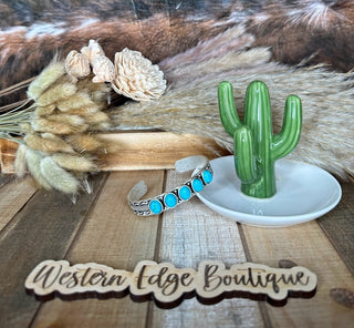 The Roux Turquoise Stone Cuff Bracelet, featuring natural stones and antique silver plate accents, rests on a wooden surface beside a small green cactus figurine on a white dish. In the background, dried flowers and pampas grass add to the aesthetic. The words "Western Edge Boutique" are written in wooden letters at the bottom.