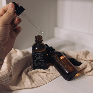 A hand holds a dropper above an open amber glass bottle labeled "Diffuser Oil 2 oz" from Western Edge Boutique, promising long-lasting scents. Another closed amber glass bottle lies on its side on a beige textured cloth next to the open bottle, set against a light background.