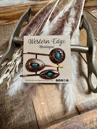 A display card from Western Edge Boutique showcases the Frankie 3 Pcs Aztec Style Hairpin Set, each featuring intricate designs and vibrant turquoise stones. The card is placed on a wooden surface with decorative feathers and hints of copper in the background.