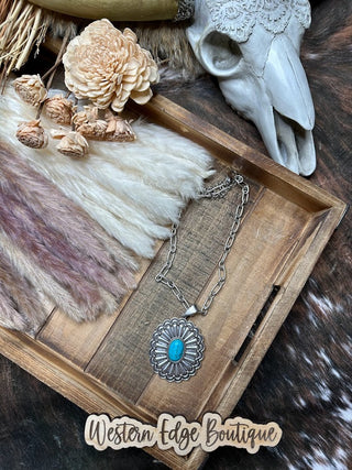 A decorative wooden tray holds the Halten Concho Turquoise Stone Necklace from Western Edge Boutique, dried flowers, and a cow skull, set on a fur background. The 15" necklace features an extender chain for adjustable jewelry. The text "Western Edge Boutique" appears at the bottom.