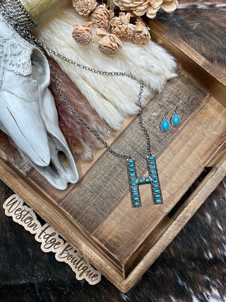 A rustic display showcases the Hannah "H" Letter Turquoise Necklace Earring Set by Western Edge Boutique, featuring a turquoise-studded "H" necklace and matching turquoise earrings. Decorative elements, including a white skull and dried flowers, are arranged on a wooden tray labeled with "Western Edge Boutique," complemented by brushed silver accents.