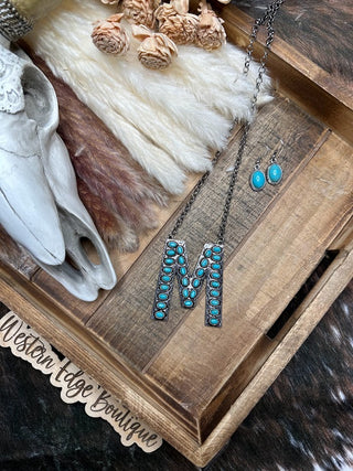 A decorative tray holds the Mallory "M" Letter Turquoise Necklace Earring Set by Western Edge Boutique, along with a textured fabric and a cow skull. Beside the tray is a wooden cutout sign that reads "Western Edge Boutique." Dried flowers in the background add charm to Ms. Mallory's display.