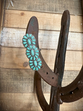 The Kieran Western Concho Stretch Ring from Western Edge Boutique, adorned with a cluster of flower-shaped turquoise stones and burnished silver detailing, is elegantly displayed hanging on a rustic sculpture crafted from repurposed horseshoes. The scene is set against a backdrop of wooden plank walls.