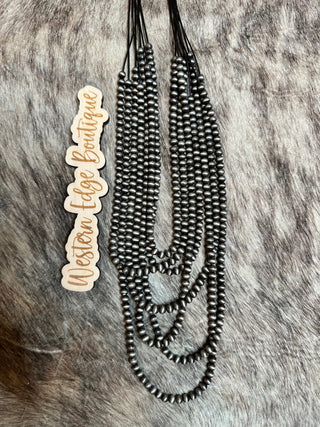 A set of Millie Navajo Pearl Layered Necklace, featuring 7 layers of black and metal beads, laid out on a textured, grayish fur background. Beside the necklaces is a wooden cutout sign that reads "Western Edge Boutique.