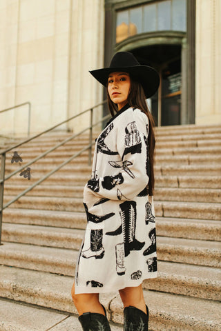 A woman in cozy boots and a 2FLY "Boots a Million" cardigan, with western-themed black and white prints, stands on stone steps. Wearing a cowboy hat, she gazes neutrally at the camera in front of large wooden doors.