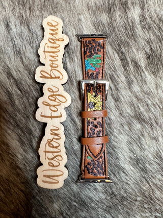 The Lasso Large Apple Watch Band from Western Edge Boutique, adorned with a floral and cactus design, lies next to a wooden sign that reads "Western Edge Boutique" on a textured, furry surface. The watch band features intricate, colorful embroidery accented with burnished silver details.