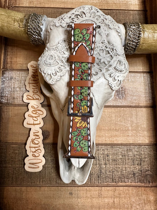 A detailed, hand-painted Josie Large Apple Watch Band with multi-color leather and green cactus and yellow flower designs is elegantly displayed on a decorative cow skull accented with lace. The background showcases wooden planks, accompanied by a wooden sign reading "Western Edge Boutique" on the left.