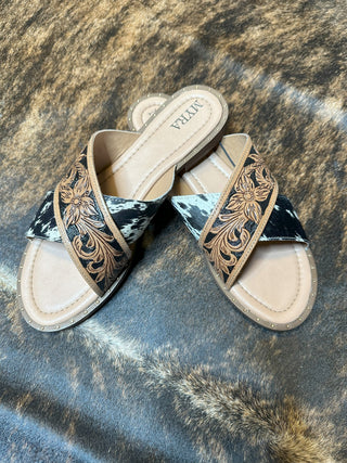 A pair of Myra Jersey Western Hand-Tooled Sandals featuring beige footbeds, black and white cowhide straps, and intricate brown floral embroidery. The classy appearance is enhanced by the durable materials, all set against a patterned brown and black background.