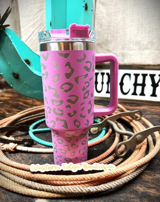 A tall, purple stainless steel tumbler with a leopard print pattern stands on a wooden surface, surrounded by coiled ropes. It's positioned in front of a vertical decor piece shaped like a teal cactus. A sign behind it reads "Western Edge Boutique.