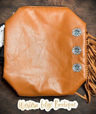 A brown faux leather handbag featuring fringe detailing on the side, adorned with three silver floral conchos and subtle embroidery, is displayed against a dark background. A label reading "2 Fly" is placed below the Kami Pistol Packin' Make-up Bag.