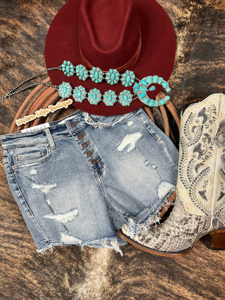 A flat lay image showcases a pair of Rebecca High Rise Button Up Shorts by VERVET by Flying Monkey, a wide-brimmed red hat, a turquoise floral necklace, a Western Edge Boutique tag, and a pair of intricately patterned cowboy boots, all laid out on a textured brown surface.