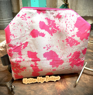 A large size, neon pink and white abstract-patterned bag named "Pink Poppin' Hide Your Crazy Travel Bag" by the brand 2 Fly features a rectangular wooden tag that reads "Western Edge Boutique." The bag is placed on a wooden surface with part of a turquoise object in the background.