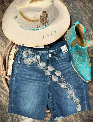 Western-themed outfit consisting of a beige cowboy hat with a cactus design, a pair of blue denim shorts, turquoise cowboy boots with intricate embroidery, and the Silver Western Concho Belt S/M from Western Edge Boutique paired perfectly with a matching necklace and lasso.