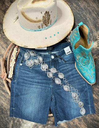 An outfit is laid out on a textured surface, featuring a beige cowboy hat with cactus designs, a pair of Judy Blue Jude Women's Side Slit Cut-off Shorts, turquoise cowboy boots with intricate stitching, and a silver concho belt. A lasso is thoughtfully positioned around the items.