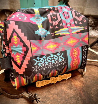 The Jacklee Neon Travel Bag by 2 Fly, featuring vibrant Southwestern patterns in shades of pink, turquoise, yellow, and black, rests on a wooden surface. Large enough to fit in your bathroom drawer, it adds a dash of style. A small sign beneath the bag reads "Western Edge Boutique." The background includes rustic wooden elements and a rope accent.