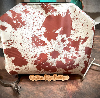 The Dusti Hide Your Crazy Travel Bag by 2 Fly is a cowhide-patterned pouch featuring brown spots on a white background, displayed on a wooden surface. This large-sized pouch includes a tag at the bottom reading "Western Edge Boutique." A turquoise item is partially visible in the background.