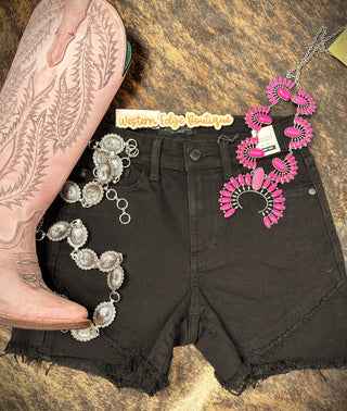 A leopard print background showcases a pair of Oakley Black Frayed Tulip Hem Shorts by Judy Blue, perfect for summer adventures, a pink cowboy boot, and two statement necklaces. One necklace features silver conchos, while the other has bold pink stones. A "Western Edge Boutique" label is placed above the comfortable shorts.