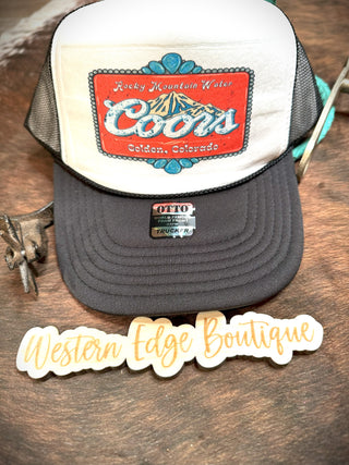 Close-up of a white and black Rocky Mountain Trucker Hat with a "Coors Golden, Colorado" patch on the front, lying on a wooden surface next to a cut-out sign that reads "Western Edge Boutique." The one-size-fits-all hat features a silver tag that says "OTTO Snapback Trucker Cap.