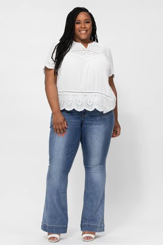 A woman stands smiling against a plain white background. She is wearing a white short-sleeve blouse with a scalloped hem and stylish Jesse Curvy Mid Rise Flare Jeans by Judy Blue. She has long braided hair and is wearing white sandals. One of her hands rests relaxed by her side while the other is placed on her thigh.