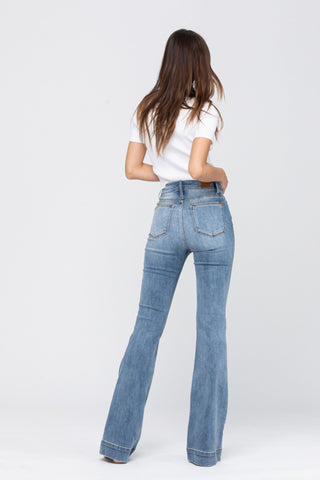 Seen from behind against a plain background, a woman with long hair wears a white short-sleeve top and Jesse Mid Rise Flare Jeans from Judy Blue in a medium wash. Her left hand rests on her hip, while her right hand adjusts her hair, embodying casual elegance with these stylish jeans.