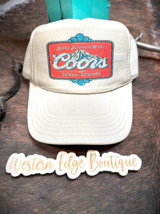 A Rocky Mountain Trucker Hat, featuring a beige design with a patch that reads "Rocky Mountain Water Coors Golden, Colorado," is displayed on a wooden surface. Below the hat, a sign reads "Western Edge Boutique" in a stylized, cursive font. This versatile hat is one-size-fits-all and perfect for any Western enthusiast.