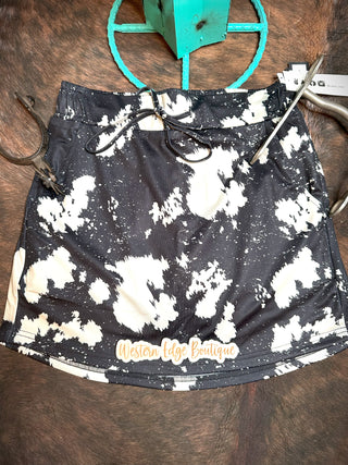 The Oh My Hide Skort from Western Edge Boutique, with its black and white speckled design, is artfully laid out on a rustic wooden surface. Made from stretchy polyester, the skort includes a drawstring waist and features a subtle logo at the hem. A turquoise decor piece is partially visible at the top of the image.