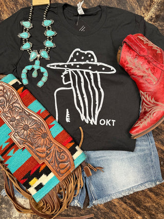 Flat lay of Western-themed items including a super soft Western Edge Boutique tee, the Harley Cowgirl Silhouette (Black), showcasing a white line drawing of a person wearing a hat. Paired with light blue denim shorts, red cowboy boots, turquoise and silver jewelry, and a multicolored woven bag with brown leather details. Perfect for the badie cowgirl look.