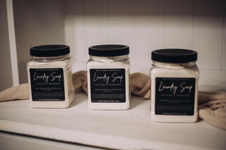 Three jars of Laundry is SO much fun when it smells this great!!! from Mav Wicks Candle Co. are displayed on a wooden shelf. The jars, perfect for deep cleaning, have black labels with white text. Two jars are positioned close to each other on the left, while one is placed slightly apart on the right.